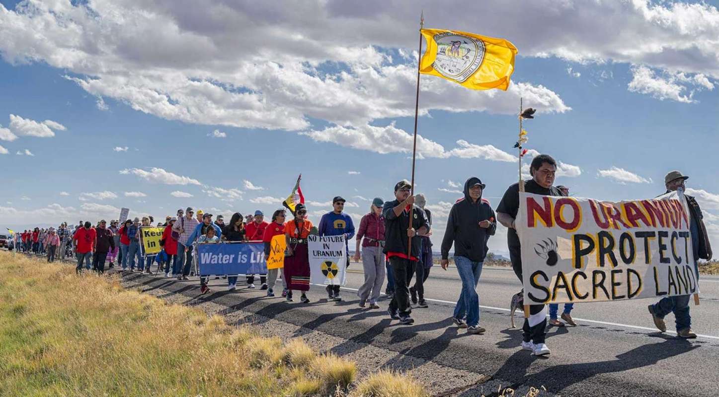 A group of people marches along a road, holding banners advocating for water protection and against uranium mining.