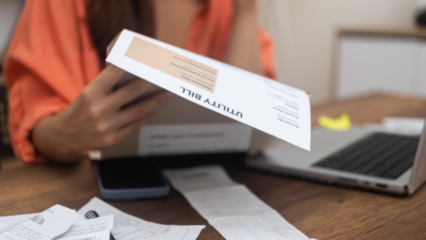 A woman examines a utility bill while surrounded by receipts and a laptop on a wooden table.