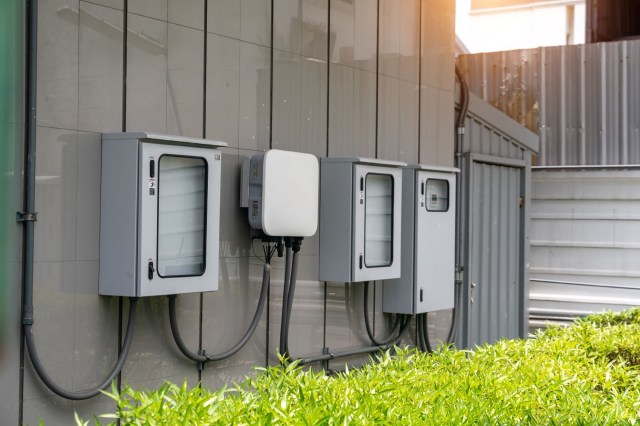 Utility boxes mounted on a wall next to a green hedge.