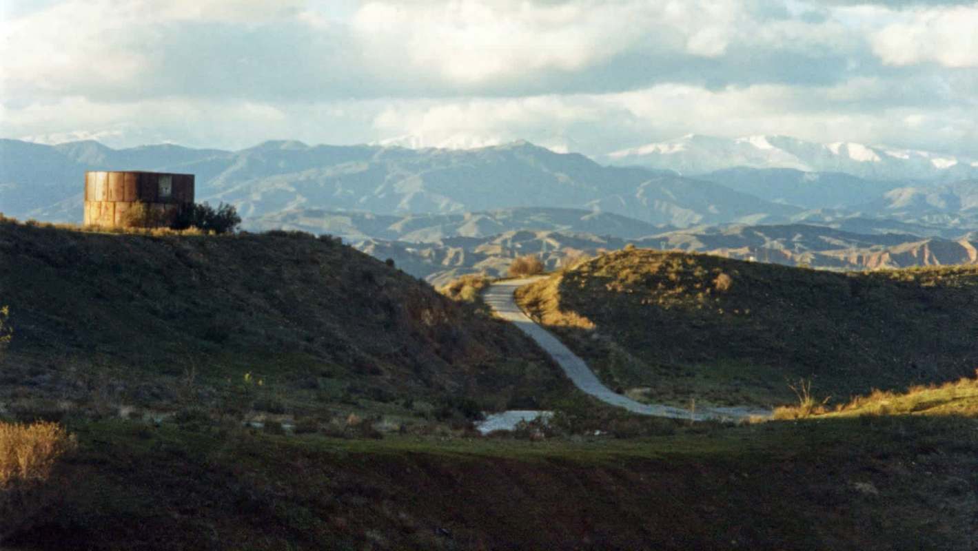 A metal structure sits atop a hill overlooking a winding road and mountainous landscape.