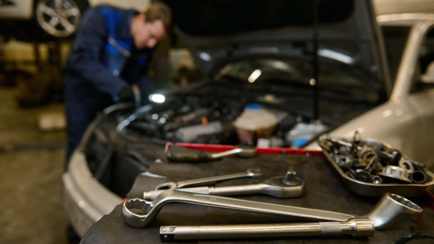 A mechanic works on a car engine while various tools are arranged on a table in the foreground.
