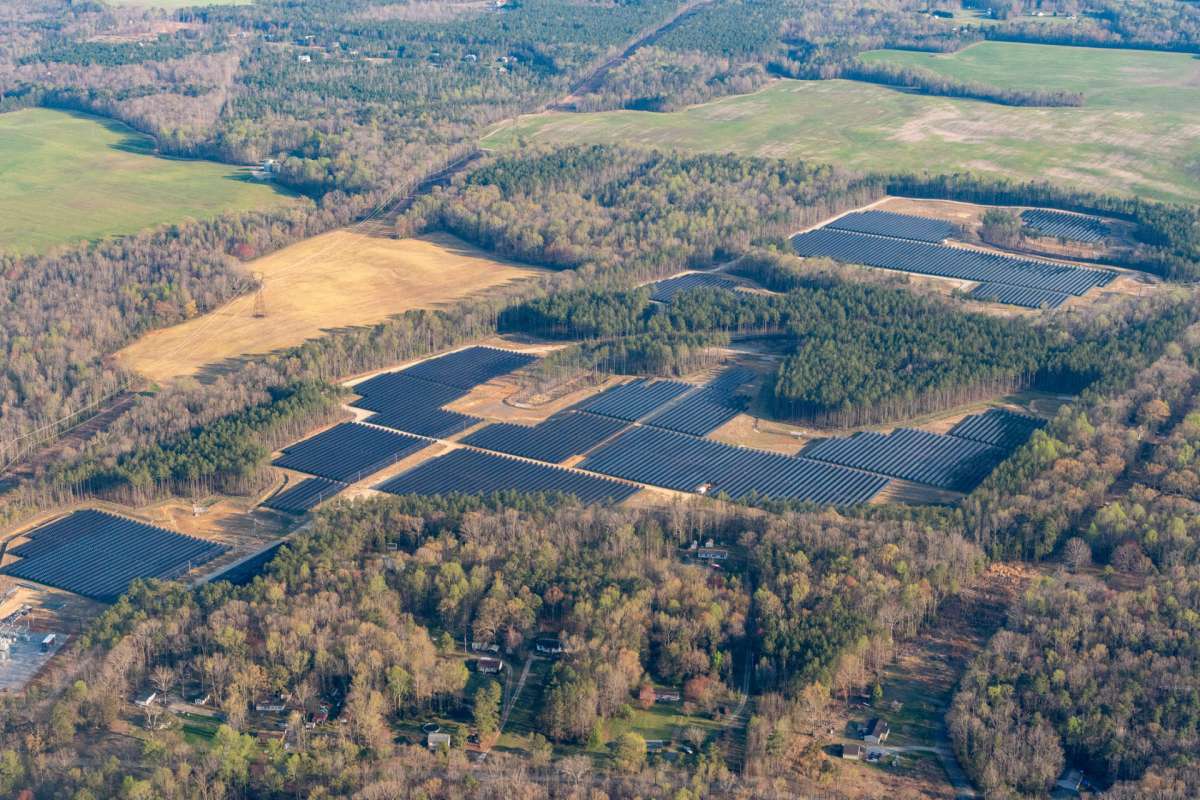 An aerial view of a large solar farm surrounded by fields and forested areas.