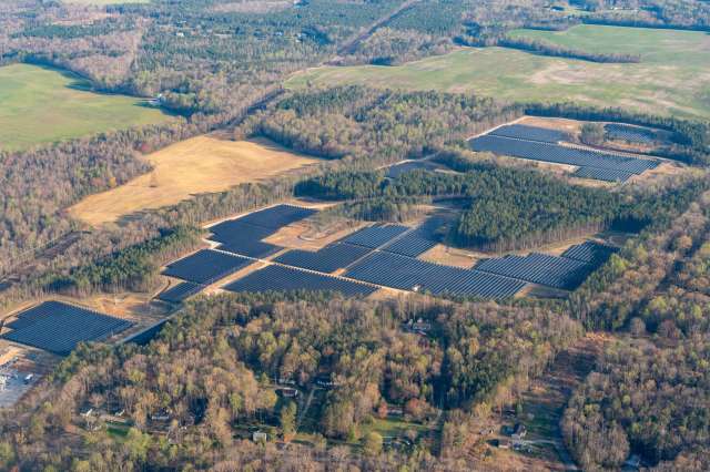 An aerial view of a large solar farm surrounded by fields and forested areas.