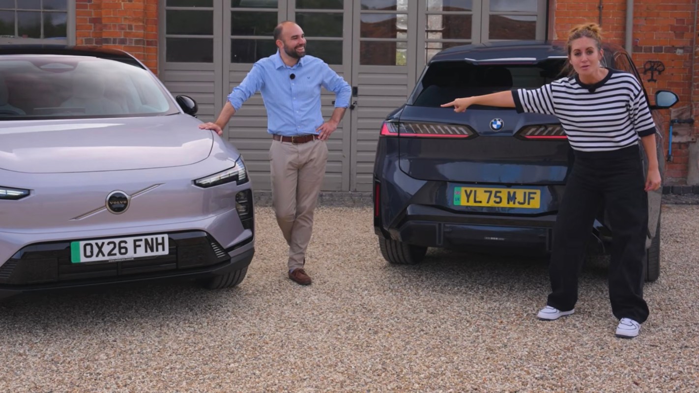 A man and a woman stand beside two parked cars, pointing and discussing features.