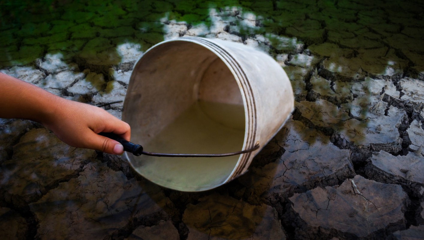 A hand holds a bucket over cracked ground with a small amount of water inside.