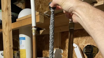 A hand holding a long, fuzzy, gray object near various pipes and tools in a workshop.