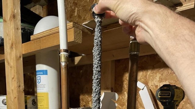 A hand holding a long, fuzzy, gray object near various pipes and tools in a workshop.