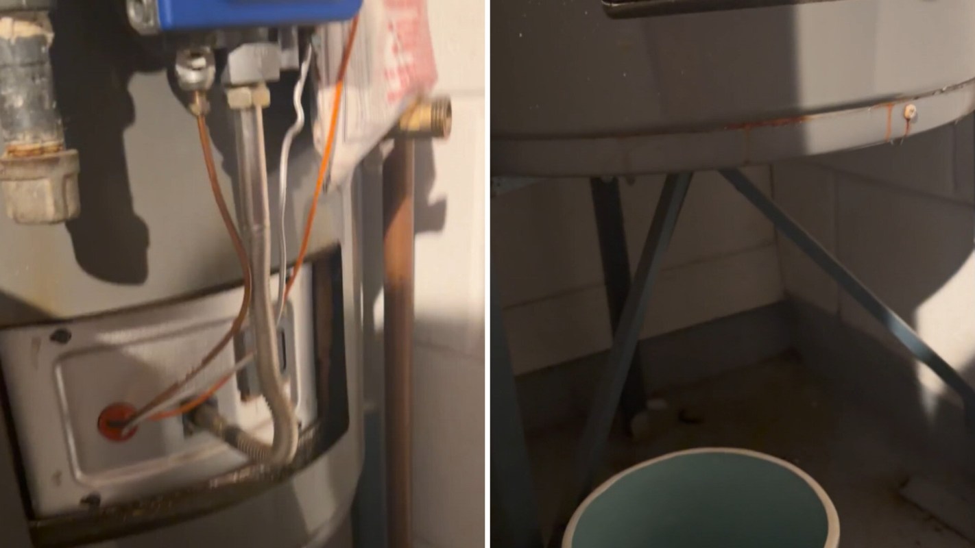A water heater with exposed pipes beside a blue bucket on a metal stand.