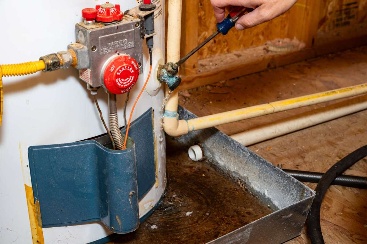 A person uses a screwdriver on a water heater valve with water pooling underneath.