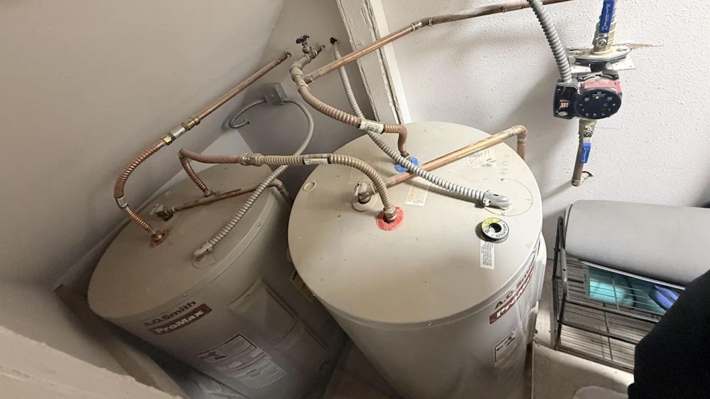 Two water heaters connected by copper pipes, with additional plumbing components against a wall.