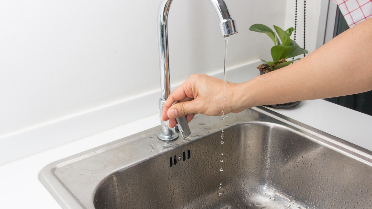 A hand adjusting a faucet with running water over a stainless steel sink.