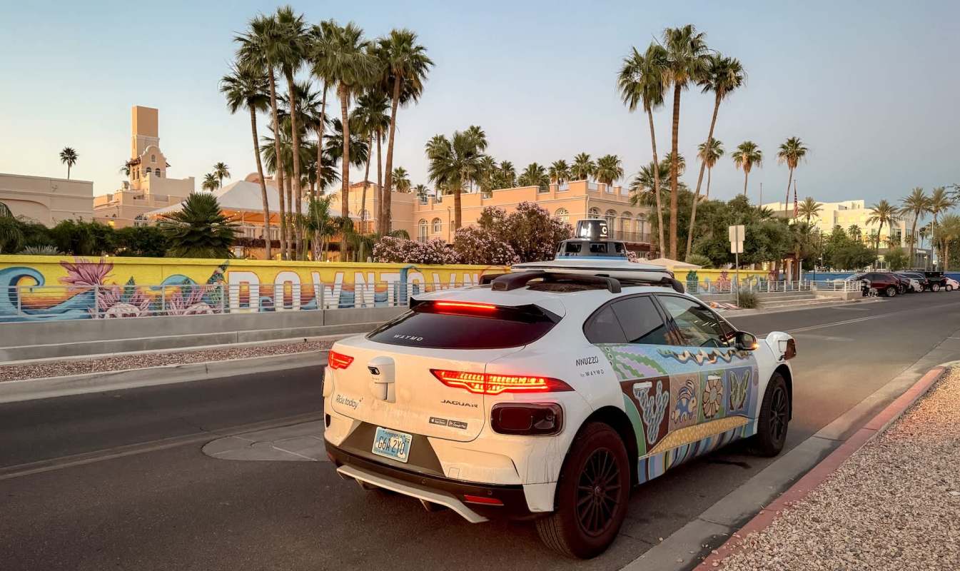 A Waymo autonomous vehicle parked near a colorful mural and palm trees in the downtown Phoenix area.