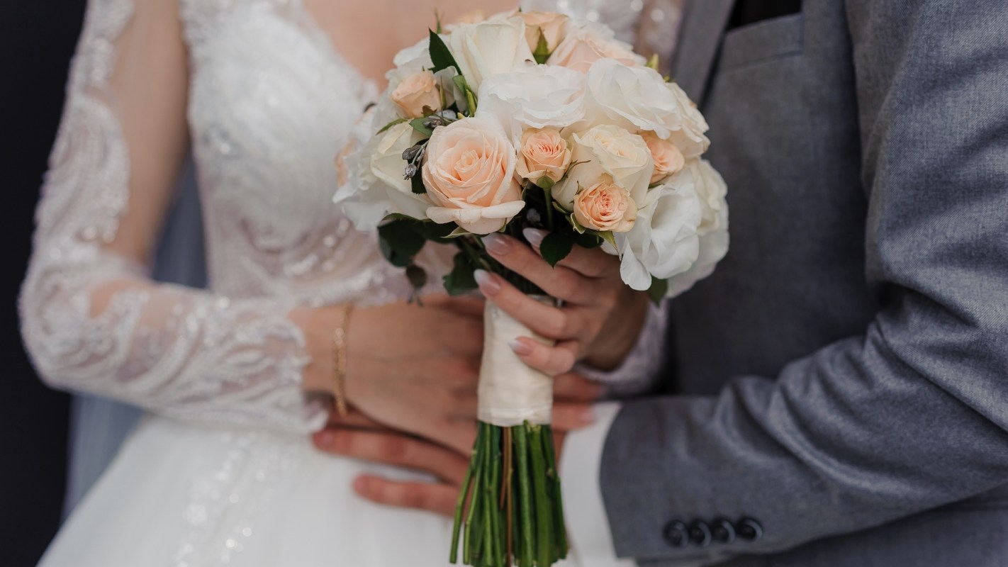 A bride holds a bouquet of roses while standing next to a person in a grey suit.