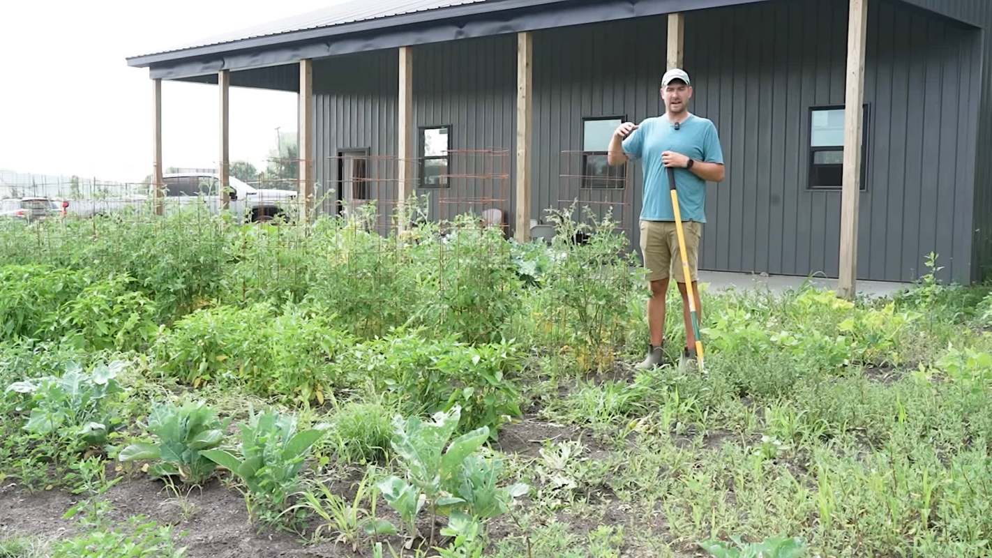 A man holding a shovel stands in a garden with various vegetables and a building in the background.