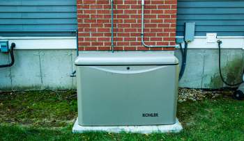 A gray generator unit positioned against a brick wall with electrical connections nearby.