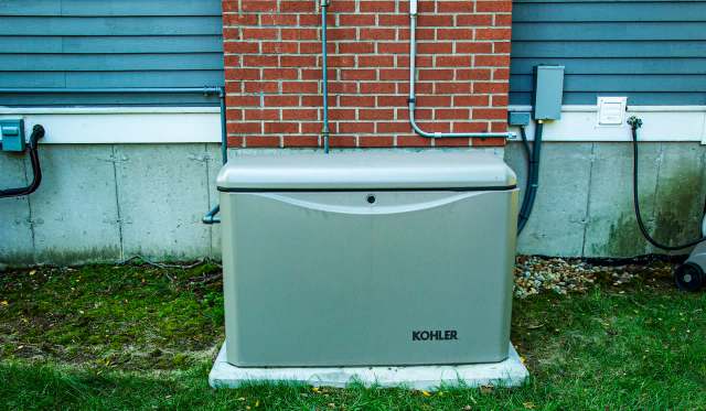 A gray generator unit positioned against a brick wall with electrical connections nearby.