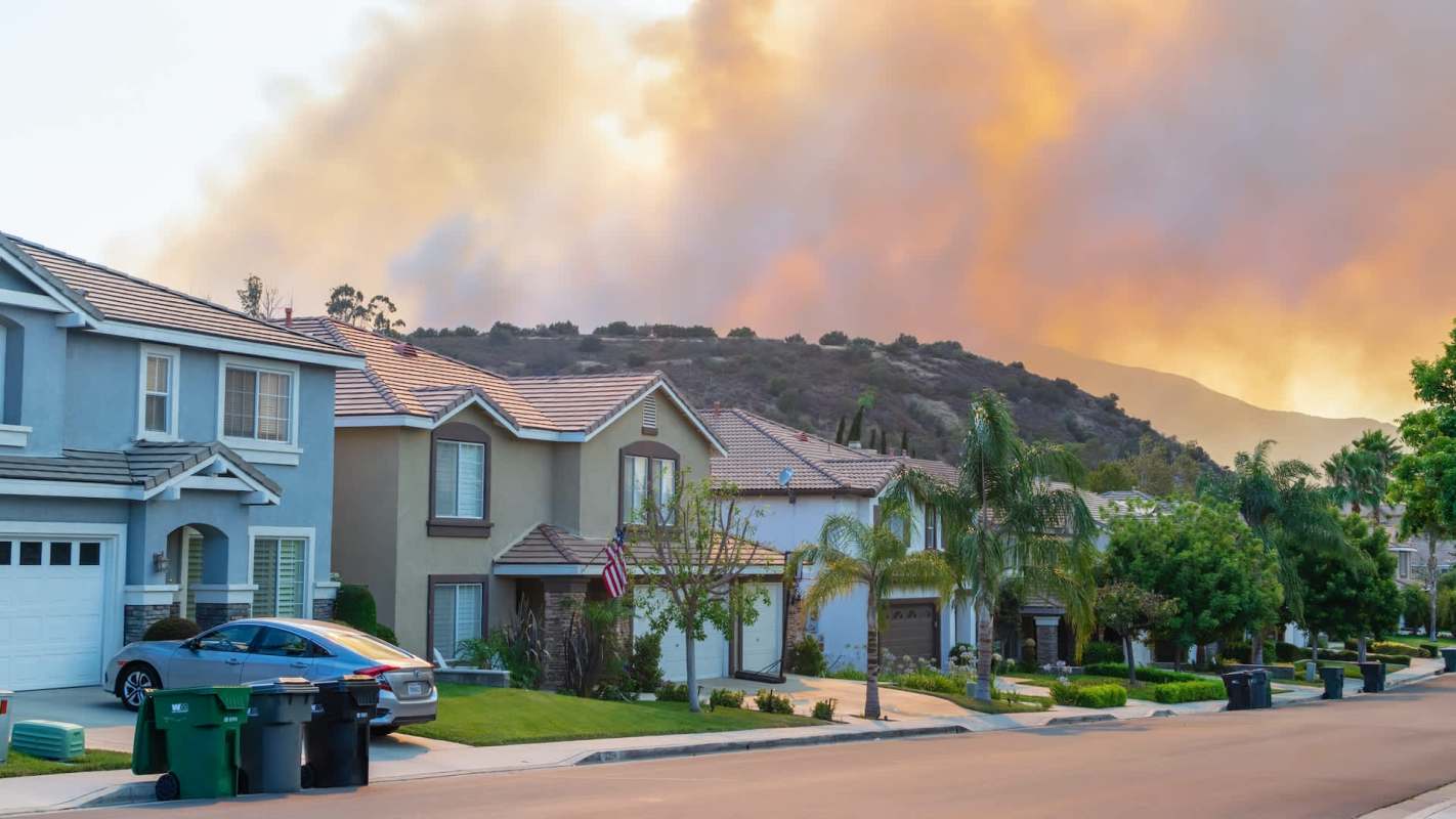 A residential neighborhood with houses, trees, and a distant smoke-filled sky.