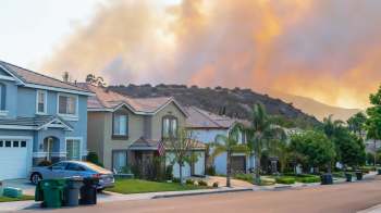 A residential neighborhood with houses, trees, and a distant smoke-filled sky.