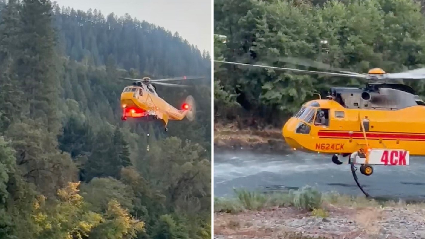 A yellow helicopter hovering above a river, equipped with firefighting gear.