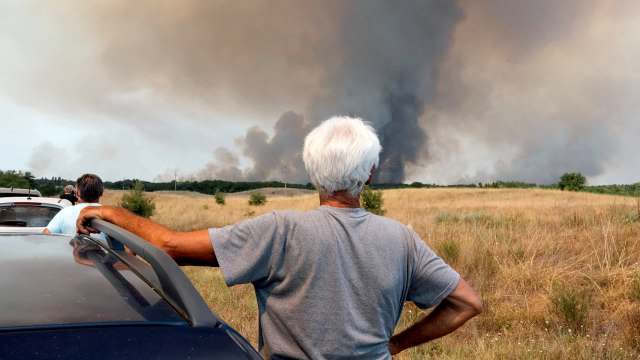 A group of people watches smoke rising from a fire in the background, with a field and trees in the foreground.