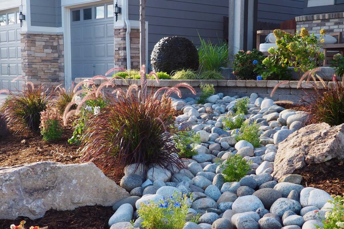 A landscaped garden featuring rocks, ornamental grasses, and various flowering plants near a house.