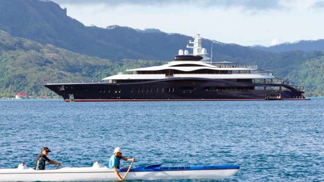 A large yacht anchored in the water with two people in a kayak nearby and mountains in the background.