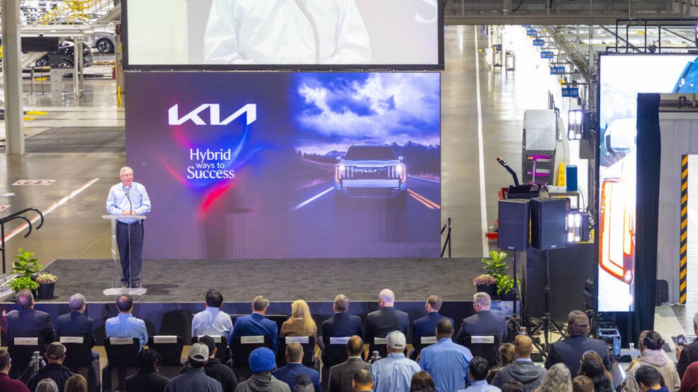 A speaker stands at a podium in front of a screen showcasing a vehicle, addressing an audience in a Kia factory setting.