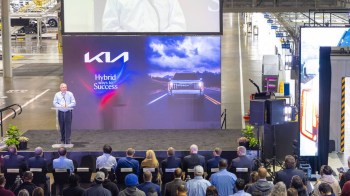 A speaker stands at a podium in front of a screen showcasing a vehicle, addressing an audience in a Kia factory setting.
