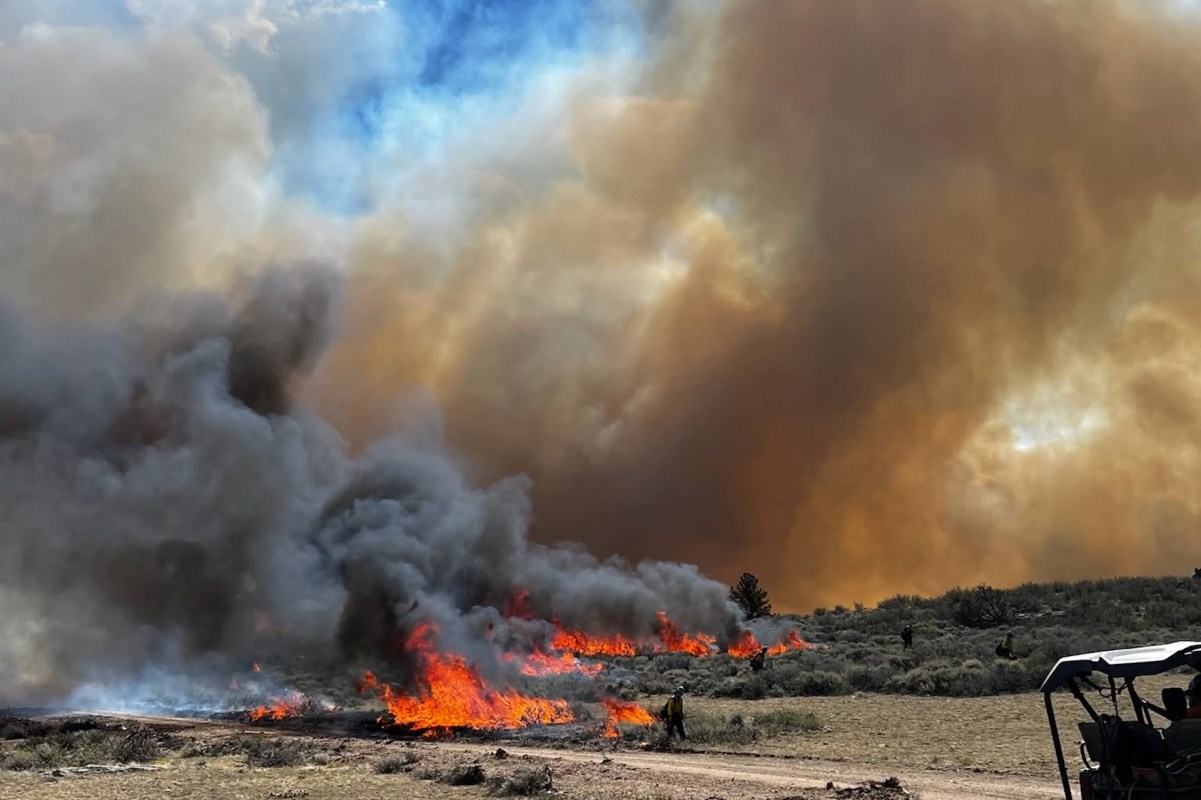 A wildfire produces flames and thick smoke in a dry landscape with a person and vehicles in the background.
