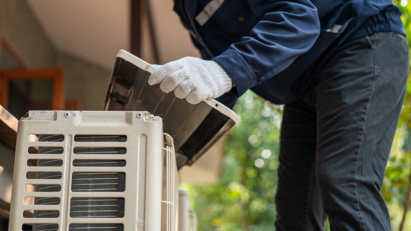 A person wearing gloves is servicing an air conditioning unit outdoors.