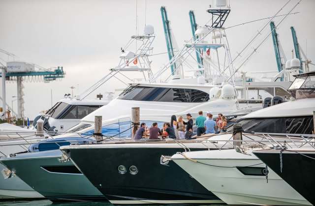 A group of people socializes on a dock among several large yachts.