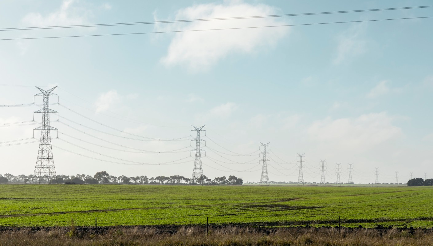 High voltage power lines stretch across a green field under a blue sky with scattered clouds.