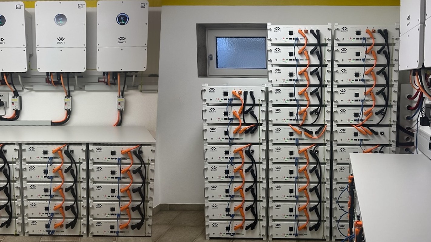 A wall of battery storage units with connected cables and inverters in a closed space.