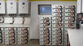 A wall of battery storage units with connected cables and inverters in a closed space.