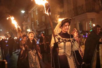 Participants in historical costumes carry torches during a nighttime event in a street.