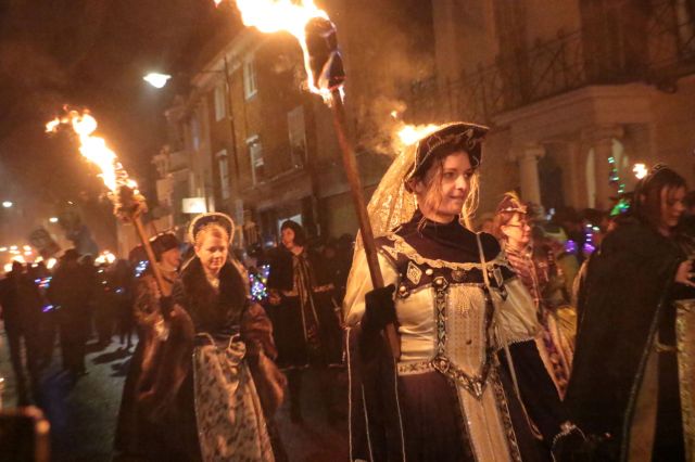 Participants in historical costumes carry torches during a nighttime event in a street.