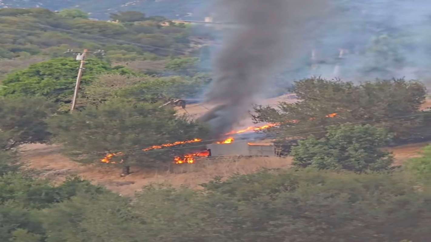 A structure engulfed in flames produces thick smoke in a wooded area.