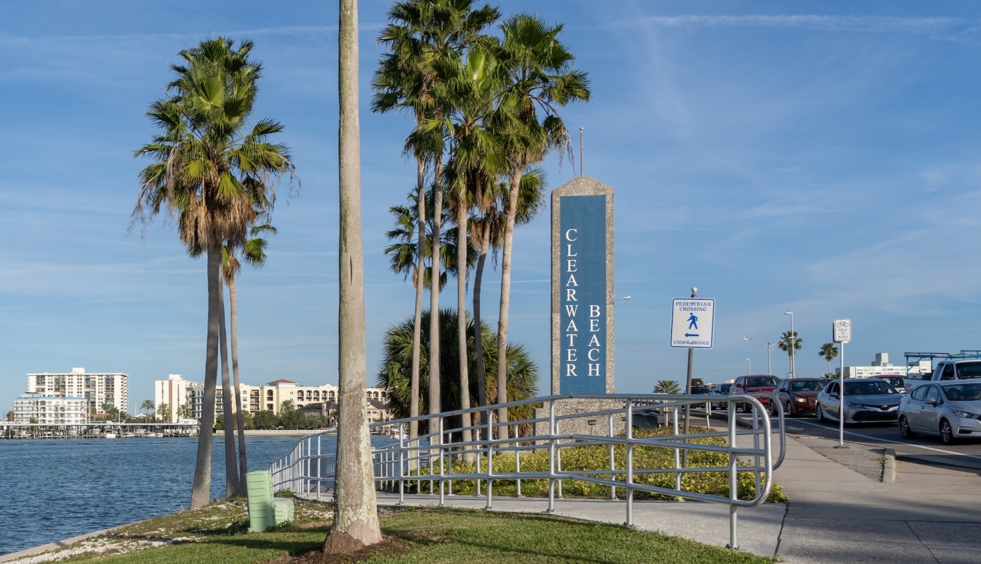A sign for Clearwater Beach is surrounded by palm trees and a waterfront pathway, with parked cars nearby.