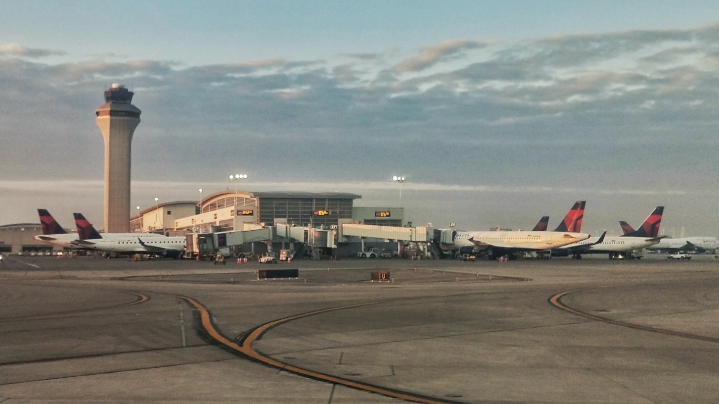 An airport terminal with multiple planes parked at the gates and a control tower in the background.