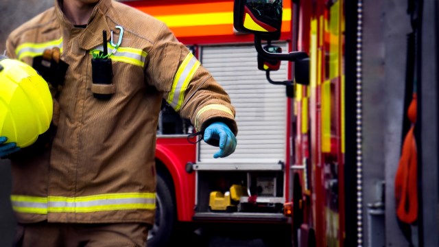 A firefighter in uniform holds a yellow helmet near a fire truck.