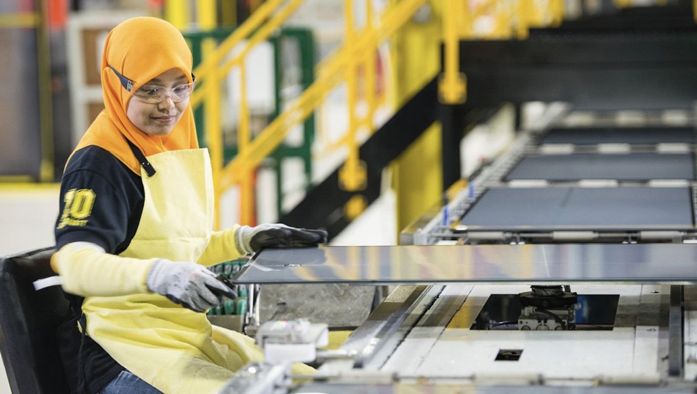 A woman in an industrial setting operates machinery while handling a large solar panel.