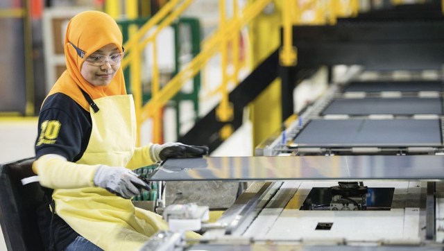 A woman in an industrial setting operates machinery while handling a large solar panel.