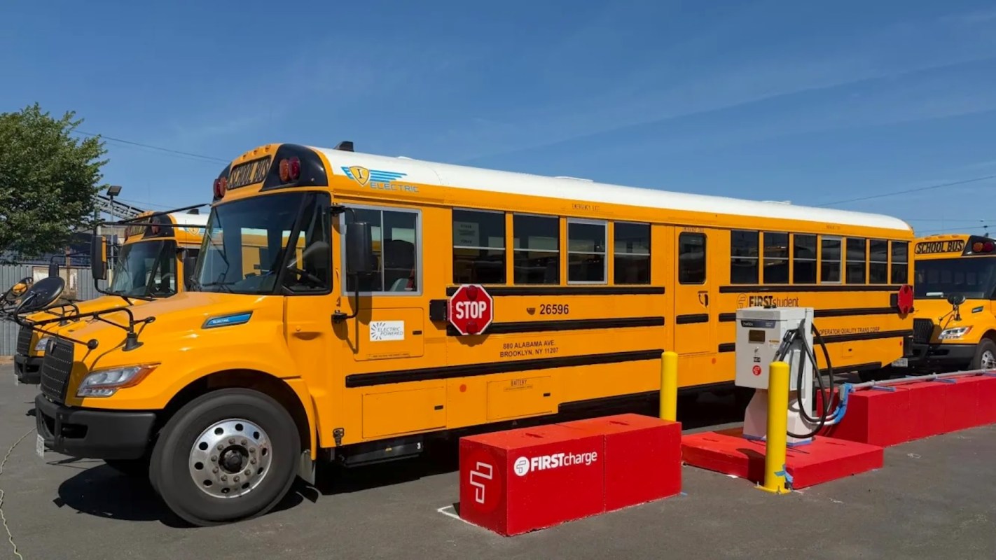 An electric school bus parked near a charging station with another bus in the background.