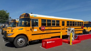 An electric school bus parked near a charging station with another bus in the background.