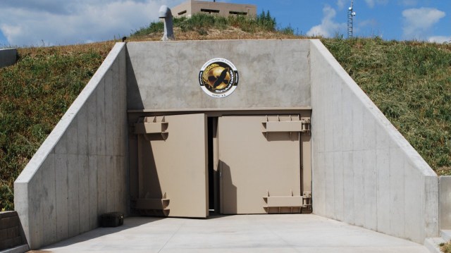 A fortified entrance with large beige doors and a logo on the wall, surrounded by grass and a hillside.