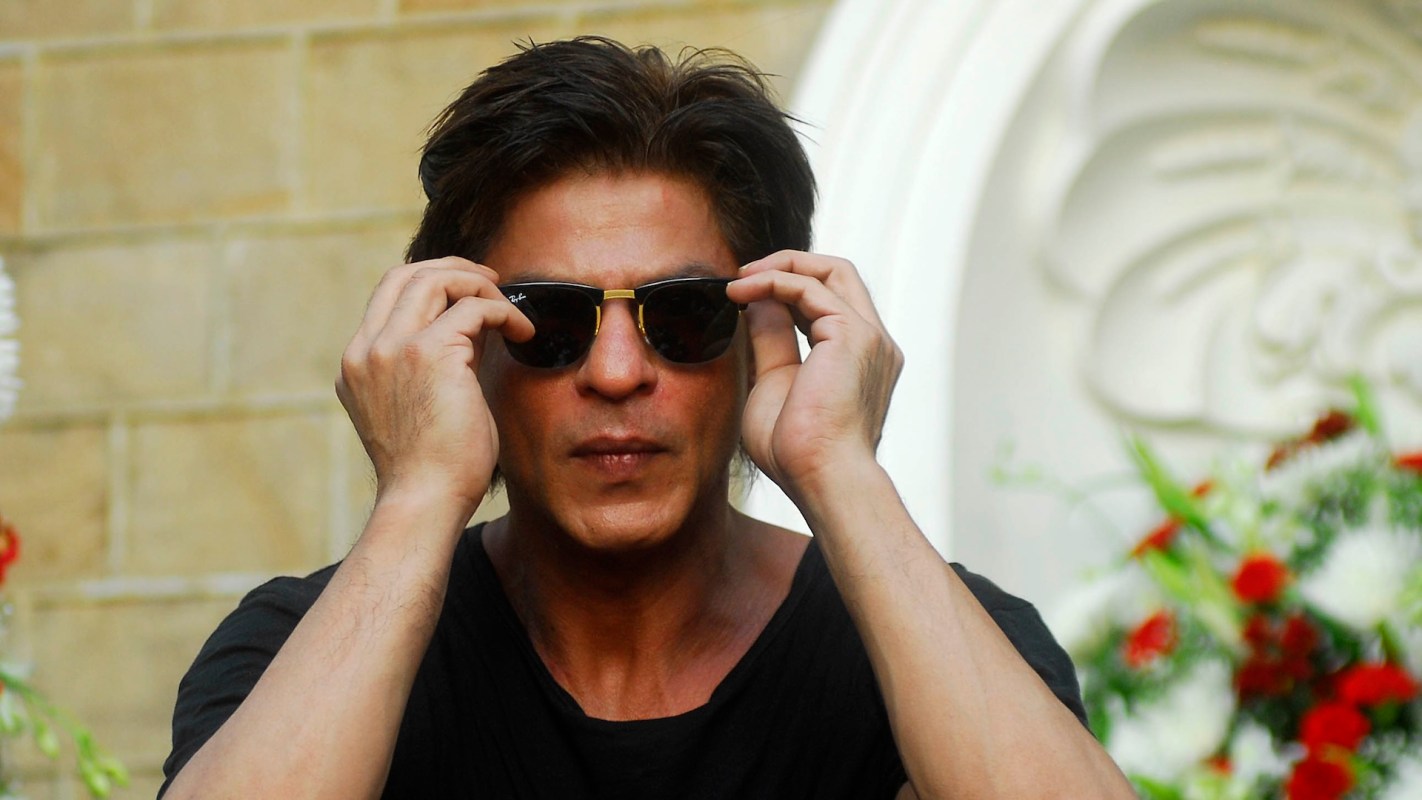 Indian actor Shah Rukh Khan adjusts his sunglasses in front of decorative flora.