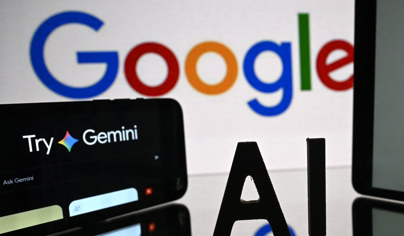 A smartphone displaying "Try Gemini" with the Google logo in the background and an "AI" letter stand.