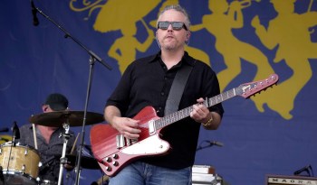 A musician plays guitar on stage with a drummer in the background.