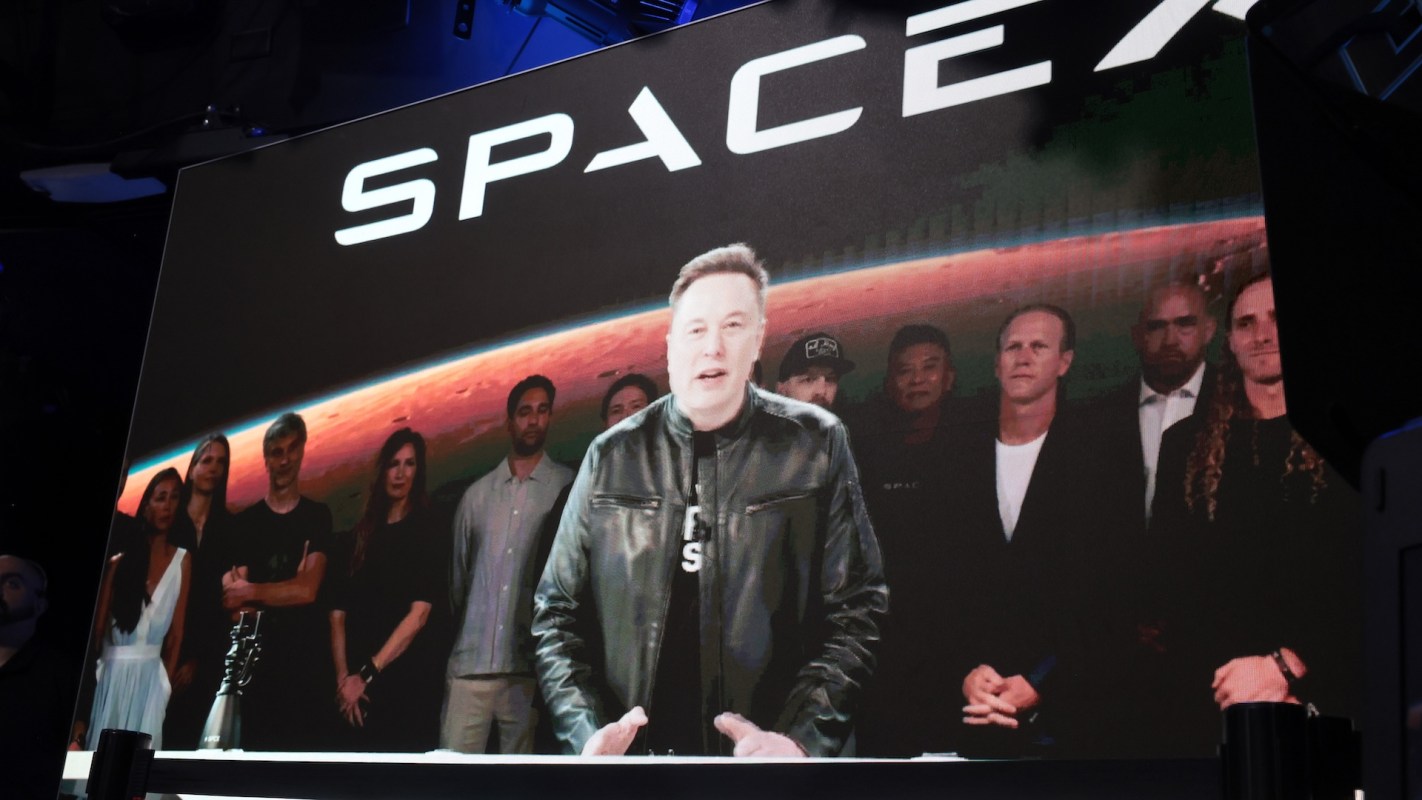 Elon Musk speaks at a Space X presentation with a group of people behind him and a space-themed backdrop featuring Mars.