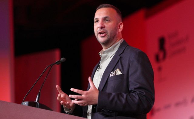 A speaker gestures while presenting at a podium during a conference.